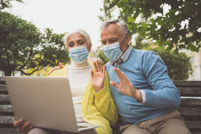 Smiling senior couple wearing mask video calling sitting at park