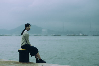 Side view of young woman sitting on bollard in sea against sky