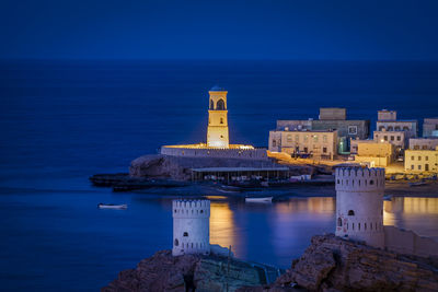 Illuminated building by sea against blue sky