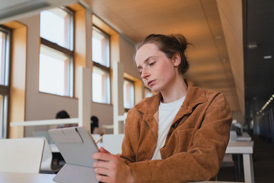 Pensive young female student with stylus leaning on hand and browsing data on tablet while sitting at table doing homework in university library