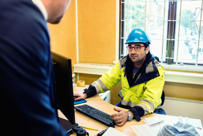 Serious construction worker in protective wear talking to man at desk