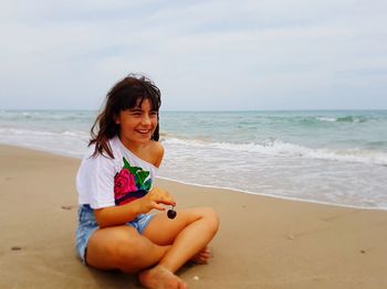 Happy teenage girl holding cherry while sitting on shore at beach against sky