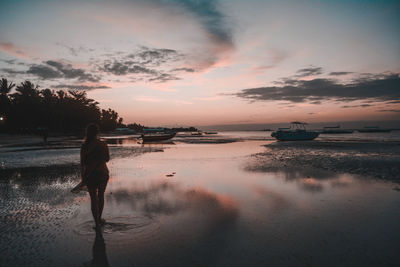 Woman walking on shore at beach against sky during sunset