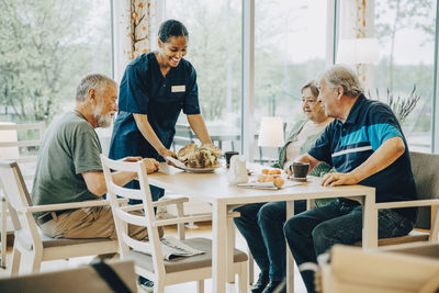 Smiling young female healthcare worker serving breakfast to senior men and woman sitting at dining table