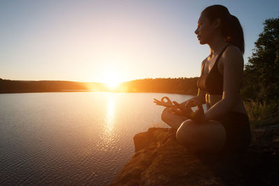 Side view of woman meditating while sitting on rock at lakeshore during sunset