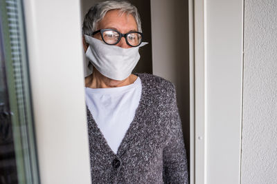 Contemplative senior adult woman standing by the window and looking out. she wears a face mask. 