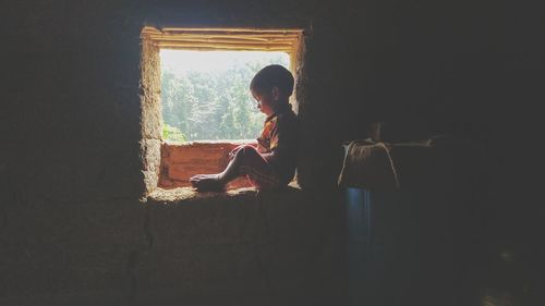 Side view of boy sitting on window sill at home