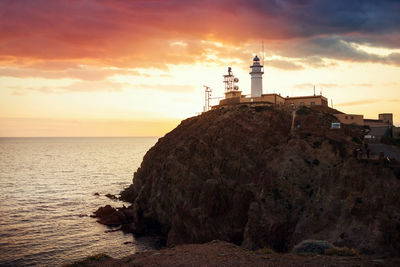 Lighthouse on rock by sea against sky during sunset