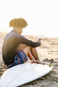 Afro surfer man consulting application on smartphone with surfboard on beach at summer sunrise