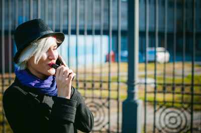 Young woman talking on mobile phone while smoking by built structure