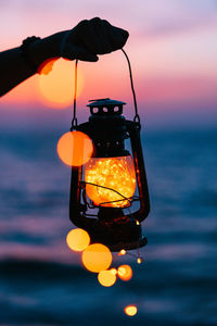 Close-up of hand holding illuminated lighting equipment against sky during sunset