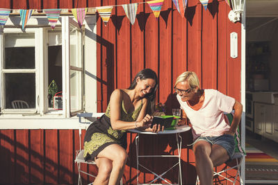 Happy woman showing mobile phone to female friend while sitting in back yard on sunny day