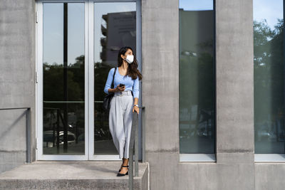 Young woman wearing protective face mask standing at entrance of building during covid-19