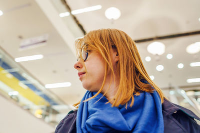 Young woman wearing eyeglasses looking away while standing against illuminated ceiling