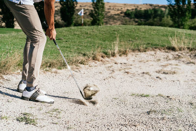 Ground level side view of crop anonymous male athlete in golf shoes hitting ball with putter in sandy hazard area of course