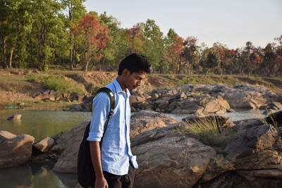 Thoughtful young man standing on rocky shore against trees