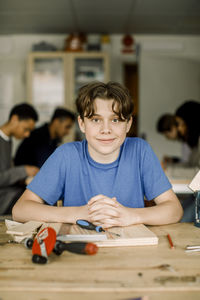 Portrait of smiling male teenage student with hands clasped at table during carpentry class