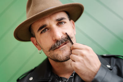 Stylish thoughtful male with earring wearing hat and leather jacket and brushing gorgeous mustache looking at camera