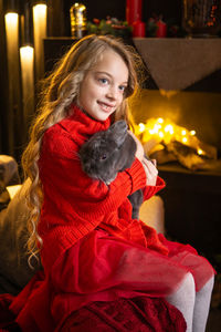 A little blonde girl with a gray rabbit in her arms next to a christmas tree decorated with garlands