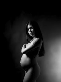 Side view portrait of naked pregnant young woman standing against black background