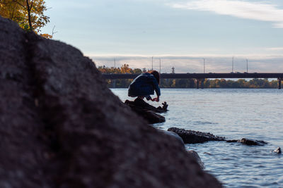 Rear view of man on rock by river against sky