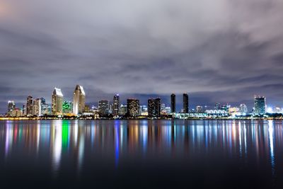 Illuminated city at waterfront against cloudy sky
