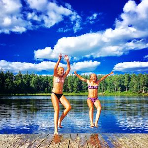 Full length of happy girls jumping in lake against sky