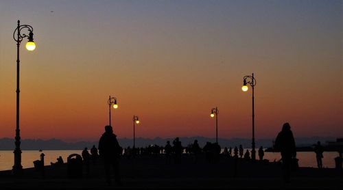 Silhouette people on illuminated pier amidst sea against orange sky during sunset