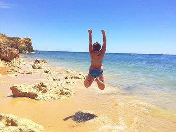 Rear view of woman in bikini jumping at beach against clear sky on sunny day