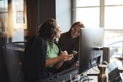 Happy businesswoman sharing smart phone with female entrepreneur sitting at desk in office