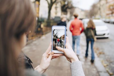Girl photographing friends through mobile phone while walking on sidewalk