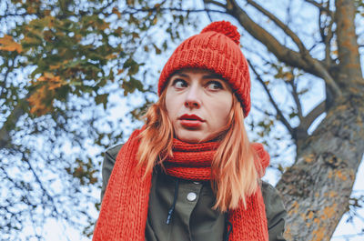 Bright cute young woman in orange warm knitted hat and scarf concerned look. blurred background