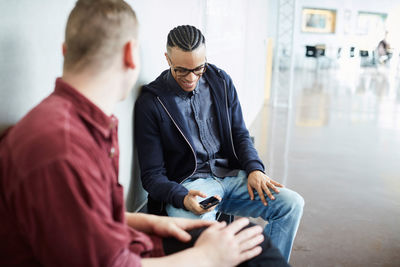 Young man using smart phone while sitting with friend on bench in corridor at university