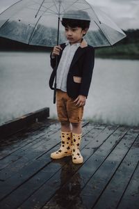 Boy with umbrella standing on pier against lake during rainfall