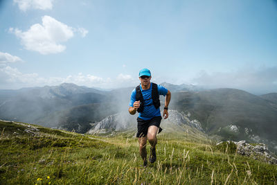 Mature sportsman trail running in the mountains on meadow against blue sky