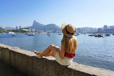 Tourist girl sitting on mureta da urca wall looking panoramic view of rio de janeiro, brazil