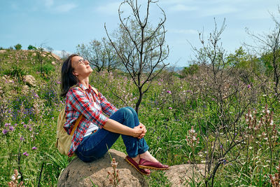 Happy carefree woman sitting on big stone in a spring meadow with beautiful field flowers.