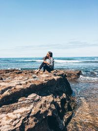 Side view of woman sitting on rock at beach against sky