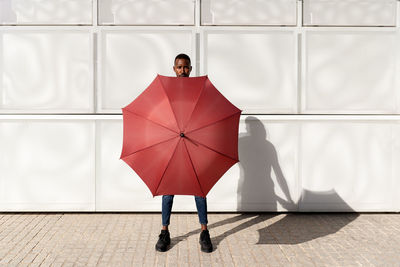 Black male standing on sidewalk in city and covering body with umbrella while looking at camera