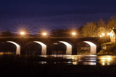 Illuminated street lights by river against sky at night