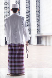 Rear view of man in traditional clothing with beaded necklace standing against wall