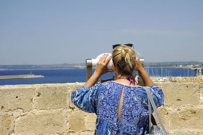 Rear view of woman looking through coin-operated binoculars against sky