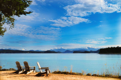 Empty chairs by lake against sky