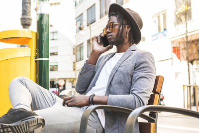 Young man talking on phone while sitting at chair against building