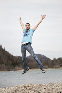 Full length portrait of man jumping against river and sky