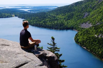 Rear view of man sitting on rock by lake against sky