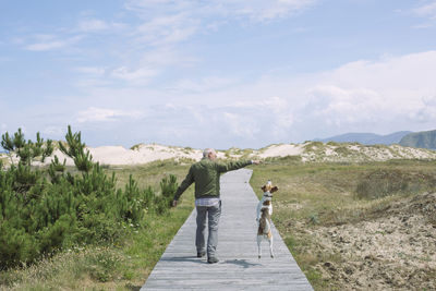 Rear view of man walking with dog on boardwalk against sky