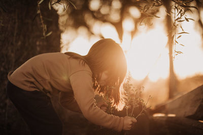 Side view of girl picking plant in park at sunset