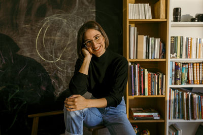 Smiling woman with hand in hair sitting against bookshelf at home