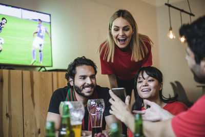 Smiling friends looking at smart phone while watching soccer in living room